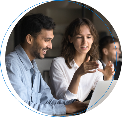 Un hombre y una mujer jóvenes en una oficina moderna trabajando juntos frente a una laptop; ella explica algo con entusiasmo mientras él mira la pantalla sonriendo.