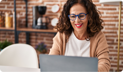 Una mujer madura con gafas de montura azul y chaqueta beige sonríe alegremente mientras mira la pantalla de su laptop en una habitación con pared de ladrillo.