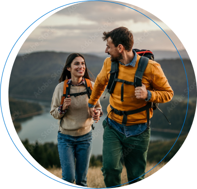 Una pareja joven con mochilas de excursión camina sonriendo por una colina frente a un lago, rodeados de un paisaje montañoso bajo la luz cálida del atardecer.