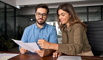 Un hombre con gafas y una mujer con camisa verde oliva revisan atentamente un documento impreso en una oficina moderna, mostrando un ambiente de trabajo colaborativo.