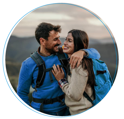 Una pareja joven con mochilas de excursión camina sonriendo por una colina frente a un lago, rodeados de un paisaje montañoso bajo la luz cálida del atardecer.